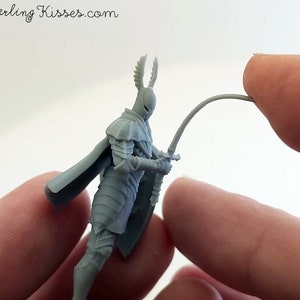 May include: A gray 3D printed miniature of a knight wearing armor and holding a sword. The knight has a cape and a helmet with horns. The miniature is being held by a hand.