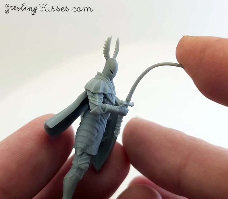 May include: A gray 3D printed miniature of a knight wearing armor and holding a sword. The knight has a cape and a helmet with horns. The miniature is being held by a hand.