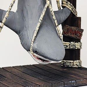 Pu&ograve; includere: Una statuetta di squalo grigio appesa a una struttura di legno con una corda. Lo squalo ha una grande bocca con denti affilati. La struttura ha un cartello con la scritta "JAWS".