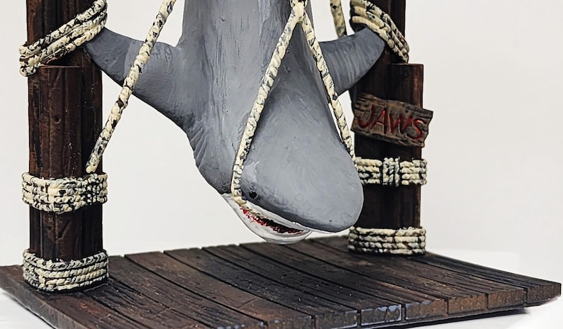 Pu&ograve; includere: Una statuetta di squalo grigio appesa a una struttura di legno con una corda. Lo squalo ha una grande bocca con denti affilati. La struttura ha un cartello con la scritta "JAWS".