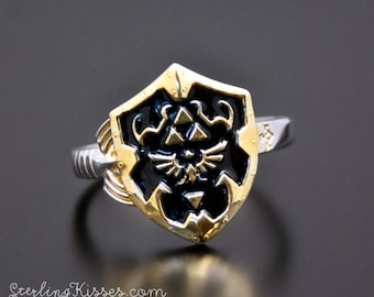 May include: A silver ring with a gold shield design featuring a black and gold Triforce symbol. The shield is raised and has a textured surface.
