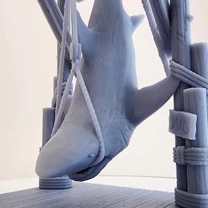 Pu&ograve; includere: Un modello 3D stampato in grigio di uno squalo appeso a una struttura in legno. Lo squalo &egrave; sospeso da corde e ha la bocca aperta. La struttura ha un cartello che recita "JAWS".