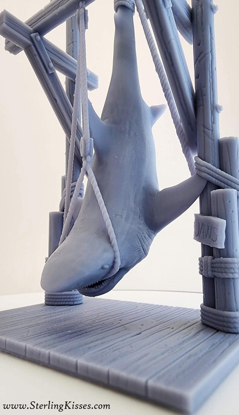 Pu&ograve; includere: Un modello 3D stampato in grigio di uno squalo appeso a una struttura in legno. Lo squalo &egrave; sospeso da corde e ha la bocca aperta. La struttura ha un cartello che recita "JAWS".