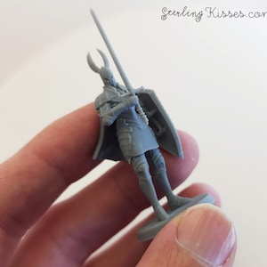 May include: A gray plastic miniature figure of a knight holding a sword and shield. The knight is wearing armor and a helmet with horns. The figure is standing on a small base.