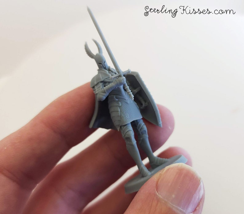 May include: A gray plastic miniature figure of a knight holding a sword and shield. The knight is wearing armor and a helmet with horns. The figure is standing on a small base.