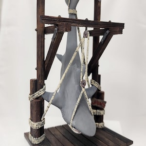 May include: A gray shark figurine hanging from a wooden structure with rope. The structure has a wooden platform and a sign that reads "Jaws".
