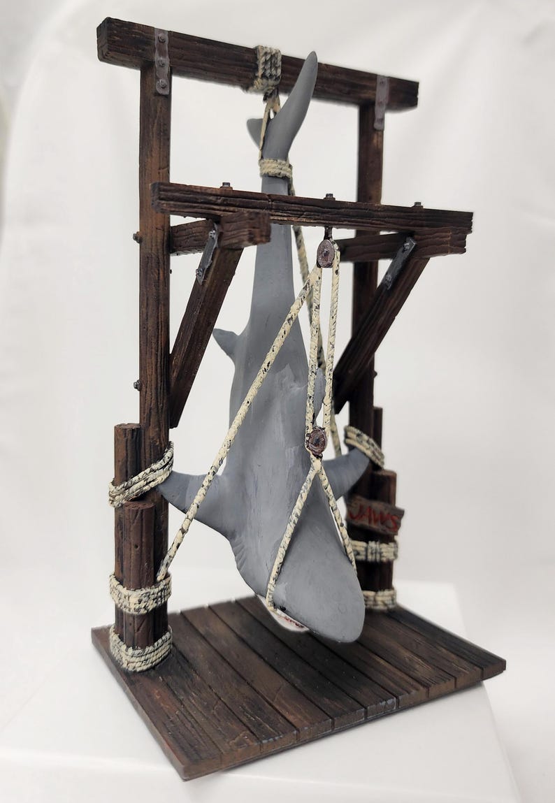 Pu&ograve; includere: Una statuetta di squalo grigio appesa a una struttura in legno con una corda. La struttura ha una piattaforma in legno e un cartello che recita "Jaws".