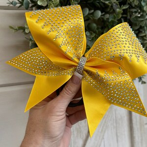 May include: A large yellow cheer bow with silver rhinestones. The bow is tied with a black elastic band.