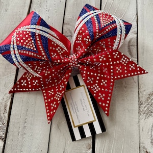 Puede incluir: Un lazo de animadora rojo, blanco y azul con purpurina y pedrería. El lazo está atado con una cinta blanca y tiene una tarjeta de visita adjunta con el texto "Bow Chik by Jenny Shaw" y "Cheer Bows & More".