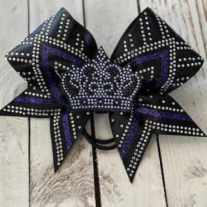 Puede incluir: Un lazo de animadora negro con un dise&ntilde;o de corona p&uacute;rpura. El lazo est&aacute; decorado con diamantes de imitaci&oacute;n iridiscentes.