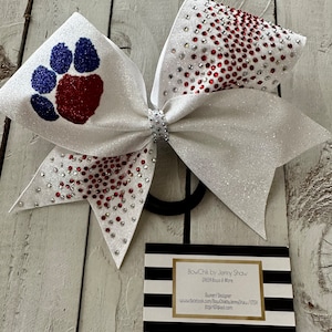Puede incluir: Lazo de animadora blanco brillante con pedrería roja y plateada. El lazo presenta un diseño de huella de pata en purpurina roja y azul. Se adjunta una banda elástica negra. En la imagen también hay una tarjeta de visita con rayas blancas y negras.