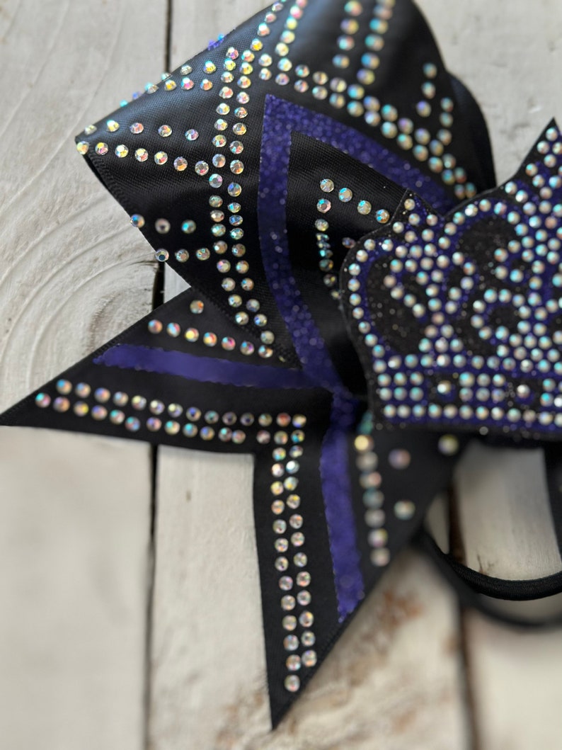 Puede incluir: Un lazo de animadora negro y morado con pedrer&iacute;a. El lazo tiene un dise&ntilde;o de estrella con rayas moradas.
