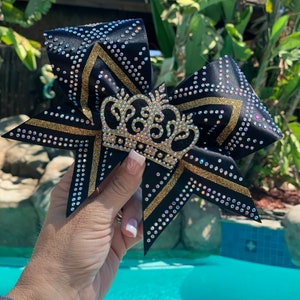 Puede incluir: Un lazo de animadora negro y dorado con un dise&ntilde;o de corona brillante. El lazo est&aacute; decorado con diamantes de imitaci&oacute;n y purpurina dorada.