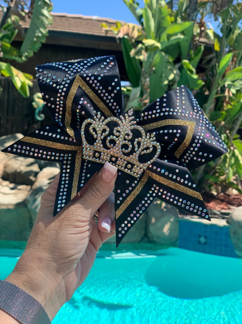 Puede incluir: Un lazo de animadora negro y dorado con un dise&ntilde;o de corona brillante. El lazo est&aacute; decorado con diamantes de imitaci&oacute;n y purpurina dorada.
