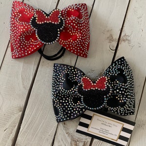 May include: Two large, sparkly hair bows with a Minnie Mouse design. The bow on the left is red with iridescent rhinestones and the bow on the right is black with iridescent rhinestones. Both bows have a black Minnie Mouse head with a red bow on top. The bows are on a white wooden surface with a business card that reads "BowChic by Janny Snow,  other stuff & more, Janny Designer, www.etsy.com/shop/BowChicbyJannySnow/  etsy.com/shop/JannySnow"