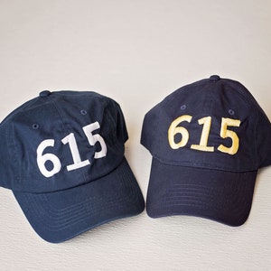 May include: Two navy blue baseball caps with the number "615" embroidered on the front. One cap has white numbers, and the other has yellow numbers. The caps are made of a soft fabric and have a curved brim.
