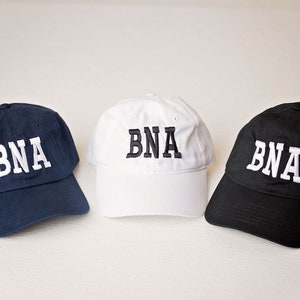 May include: Three baseball caps, one navy blue, one white, and one black, all with the letters "BNA" embroidered in white on the front.