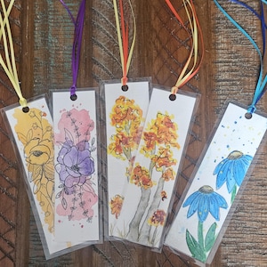 May include: Five handmade bookmarks with watercolor floral designs. The bookmarks feature a variety of flowers, including purple, blue, yellow, and orange. Each bookmark is attached to a ribbon.