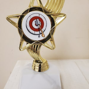 May include: A gold-colored trophy shaped like a star with a white base. The star has a red and white bullseye target with an axe in the center. The trophy is for an axe throwing competition.
