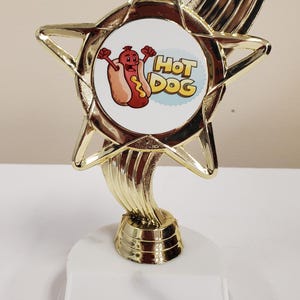 May include: A gold-colored plastic trophy featuring a star-shaped top with a circular insert. The insert displays a cartoon hot dog character with arms raised and the text "HOT DOG" in yellow letters. The trophy stands on a white rectangular base.