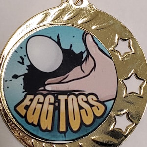 May include: Gold-colored medal with a blue, yellow, and black cartoon image of a hand holding an egg. The text "EGG TOSS" is printed on the medal. The medal has three cut-out stars around the edge.
