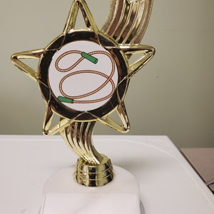 May include: Gold star-shaped trophy with a white circular plaque featuring a red jump rope and green handles. The base of the trophy is white with a blank plaque for engraving.
