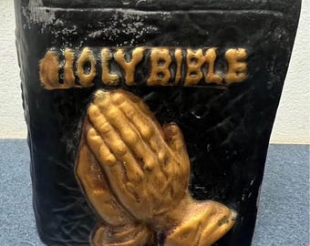 May include: Black ceramic planter shaped like a book with gold lettering that reads "HOLY BIBLE". The front of the planter features a pair of gold praying hands.