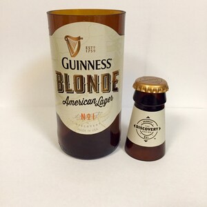 Może przedstawiać: Dwie szklane butelki, jedna większa od drugiej. Większa butelka jest brązowa i ma etykietę z napisem "Guinness Blonde American Lager No.1 Discovery Series Made in USA". Mniejsza butelka jest brązowa i ma etykietę z napisem "Guinness The Discovery Series No.1".