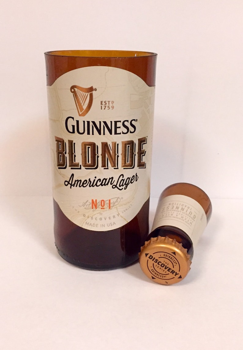 Może przedstawiać: Brązowa szklana butelka z białą etykietą, na kt&oacute;rej widnieje napis "GUINNESS BLONDE American Lager" oraz "NO.1 THE DISCOVERY SERIES MADE IN USA". Butelka znajduje się obok złotego kapsla z napisem "GUINNESS THE DISCOVERY SERIES".