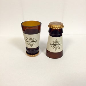 Puede incluir: Dos vasos de chupito de vidrio marr&oacute;n con tapas doradas. Los vasos est&aacute;n etiquetados con "The Discovery Series" y "No. 1".