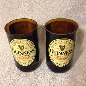 May include: Two black glass mugs with a brown top, each with a Guinness label that reads "Traditionally Brewed 17 59 Guinness Extra Stout St. James's Gate Dublin 12 fl oz."