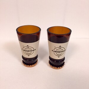 Puede incluir: Dos vasos de chupito de vidrio marr&oacute;n con tapas de botella doradas. Los vasos est&aacute;n decorados con una etiqueta blanca que dice "The Discovery Series No. 1".