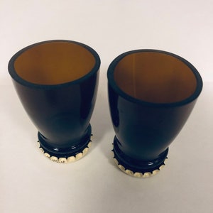 May include: Two black shot glasses with brown interiors. The glasses are made from recycled glass bottles and have bottle caps as bases.