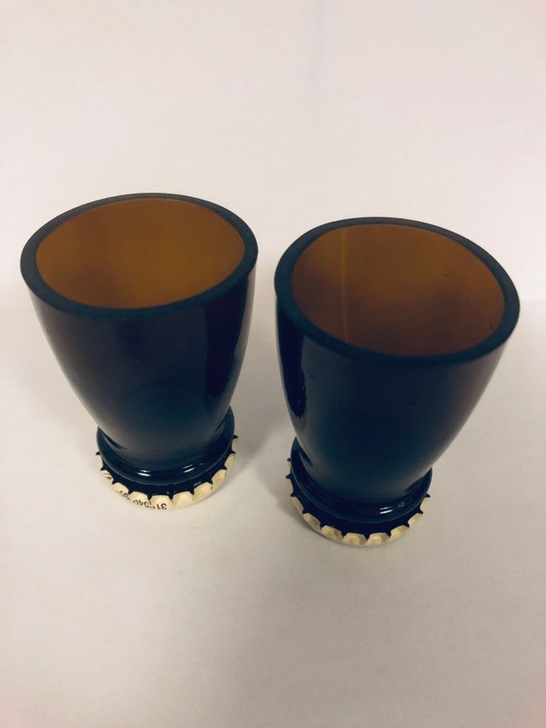 May include: Two black shot glasses with brown interiors. The glasses are made from recycled glass bottles and have bottle caps as bases.