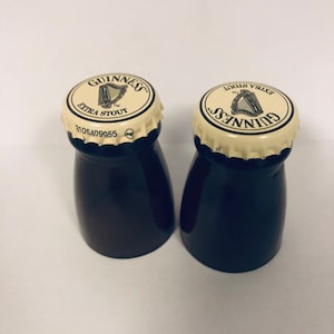 May include: Two black glass salt and pepper shakers with white lids. The lids have the Guinness logo and text "Extra Stout" printed on them.