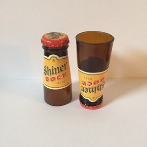 Pode incluir: Duas garrafas de vidro em miniatura, uma cheia de um l&iacute;quido castanho e etiquetada "Shiner Bock Beer", a outra vazia e etiquetada "Shiner Bock Beer".