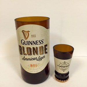 Może przedstawiać: Dwie szklane butelki, jedna wysoka, a druga niska, obie z brązowego szkła i złotymi etykietami. Wysoka butelka to butelka Guinness Blonde American Lager. Niska butelka to butelka Guinness Discovery No. 1.