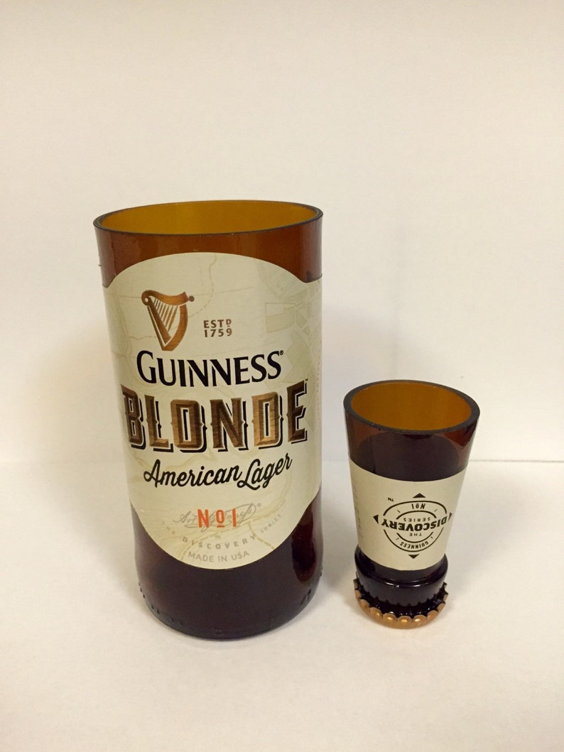 Może przedstawiać: Dwie szklane butelki, jedna wysoka, a druga niska, obie z brązowego szkła i złotymi etykietami. Wysoka butelka to butelka Guinness Blonde American Lager. Niska butelka to butelka Guinness Discovery No. 1.