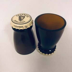 May include: Two black Guinness beer bottle shot glasses. The top of one shot glass has a Guinness Extra Stout cap with the Guinness harp logo.