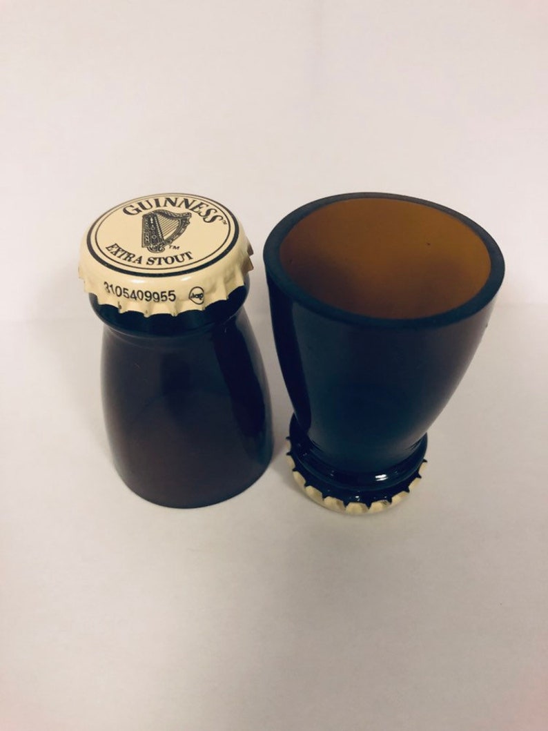 May include: Two black Guinness beer bottle shot glasses. The top of one shot glass has a Guinness Extra Stout cap with the Guinness harp logo.