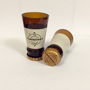 Puede incluir: Dos vasos de chupito de vidrio marr&oacute;n con tapas de botella doradas. Las tapas de botella tienen el texto "The Discovery Series" impreso.