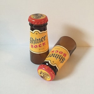 Pode incluir: Duas garrafas de vidro em miniatura de cerveja Shiner Bock. As garrafas s&atilde;o de vidro castanho com um r&oacute;tulo amarelo e uma tampa vermelha. O r&oacute;tulo apresenta uma estrela e o texto "Shiner Bock Beer".