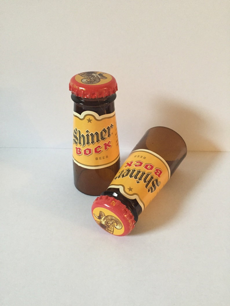 Pode incluir: Duas garrafas de vidro em miniatura de cerveja Shiner Bock. As garrafas s&atilde;o de vidro castanho com um r&oacute;tulo amarelo e uma tampa vermelha. O r&oacute;tulo apresenta uma estrela e o texto "Shiner Bock Beer".