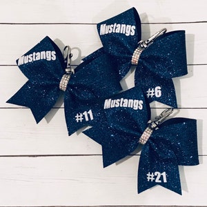 Può includere: Tre fiocchi di cheerleader blu navy con glitter, testo bianco e strass. I fiocchi hanno la parola "Mustangs" e un numero su ogni fiocco. I numeri sono #6, #11 e #21.