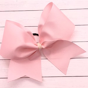 May include: A large, pale pink hair bow made from grosgrain ribbon. The bow is secured with a black elastic band and features a small, sparkly embellishment at the centre. A classic hair accessory, suitable for cheerleading or general use.