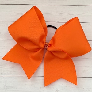 Op de afbeelding: Een grote oranje cheer bow met een glittercentrum. De strik is vastgebonden met een zwart elastiek.