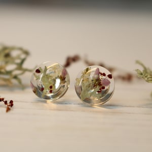 May include: A pair of clear resin earrings featuring pressed flowers and leaves. The earrings are round and have a delicate, natural look.
