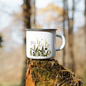 Puede incluir: Taza de esmalte blanco con un diseño floral que incluye helechos, setas y flores silvestres. La taza está sentada sobre un tocón de árbol en un entorno boscoso.