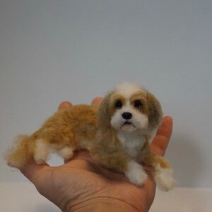 May include: A small, needle-felted dog figurine with brown and white fur. The dog is lying on its belly with its head raised and looking directly at the camera.