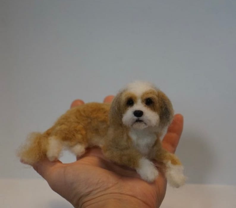 May include: A small, needle-felted dog figurine with brown and white fur. The dog is lying on its belly with its head raised and looking directly at the camera.